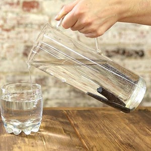 May include: A glass pitcher filled with water and vanilla beans is being poured into a glass. The pitcher and glass are on a wooden table.