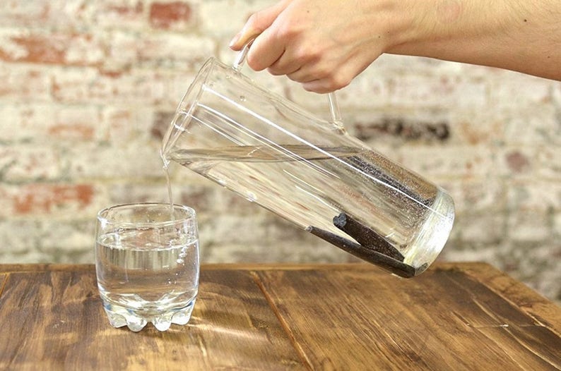 May include: A glass pitcher filled with water and vanilla beans is being poured into a glass. The pitcher and glass are on a wooden table.