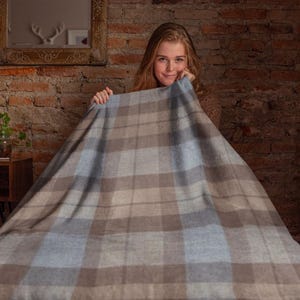 May include: A large, plaid blanket in shades of blue, gray, and brown is held up by a person. The blanket has a classic check pattern. The background features a brick wall, a wooden side table, and a mirror.