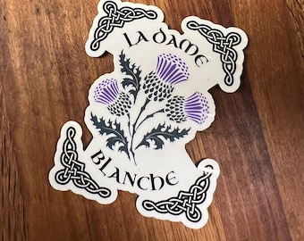 Könnte beinhalten: Ein weißer Aufkleber mit schwarzen keltischen Knotendesigns und dem Text "La Dame Blanche" in schwarzen Buchstaben. Der Aufkleber zeigt eine stilisierte Distelblume in Lila und Grau.