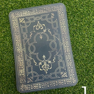 Puede incluir: Una tarjeta azul marino con un diseño dorado ornamentado. La tarjeta rectangular tiene un borde decorativo y motivos florales. El número "1" es visible en la esquina inferior derecha.