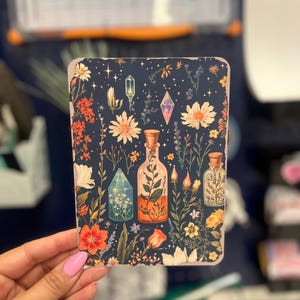 May include: A dark blue background with a floral pattern featuring various flowers, leaves, and crystals. Two glass bottles with cork stoppers are filled with colorful liquids and surrounded by the floral pattern.