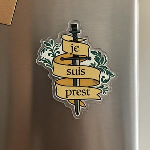 May include: A sticker featuring a sword piercing a banner with the French words "je suis prest" in a serif font. The banner is tan with a decorative green floral design around the edges.