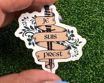 Könnte beinhalten: Ein Aufkleber mit einem Schwert und einem Banner, auf dem "Je suis prest" auf Französisch steht. Das Schwert ist schwarz und das Banner ist braun mit grünen floralen Akzenten.