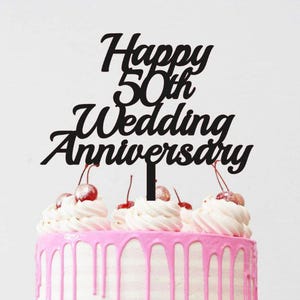Op de afbeelding: Een witte taart met roze glazuur druppels en kleurrijke sprinkles, gegarneerd met slagroom en kersen. Een zwarte taarttopper leest "Happy 50th Wedding Anniversary". De taart staat op een roze taartstandaard.