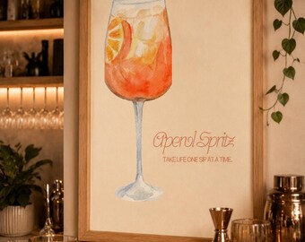 May include: A framed watercolor print featuring an orange Aperol Spritz cocktail in a stemmed glass with an orange slice garnish. The text reads Aperol Spritz and TAKE LIFE ONE SIP AT A TIME in a light orange font on a cream-colored background.
