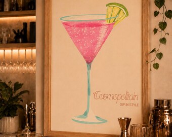 May include: A framed wall art print featuring a stylized illustration of a pink Cosmopolitan cocktail in a martini glass with a lime wedge garnish. The text "Cosmopolitan" and "SIP IN STYLE" is printed in a script and sans-serif font below the glass.