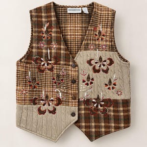 May include: A sleeveless vest featuring a patchwork design of brown plaid fabric and beige cable-knit panels. The garment is adorned with embroidered floral motifs in shades of red, white, and dark brown. It has a button-front closure and a label reading DRESS BARN.
