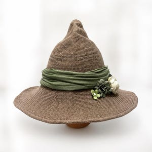 Puede incluir: Un sombrero puntiagudo de color marrón con ala ancha y una textura de punto. El sombrero presenta una banda de tela verde oliva envuelta alrededor de la base, adornada con un grupo de pequeñas flores artificiales en tonos crema y verde salvia.
