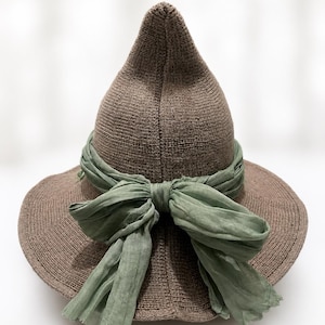 May include: A pointed brown knit hat featuring a wide brim and a sage green fabric ribbon tied into a bow around the base of the crown. The hat is constructed from a textured, solid-colored knit material with a distinct conical shape.