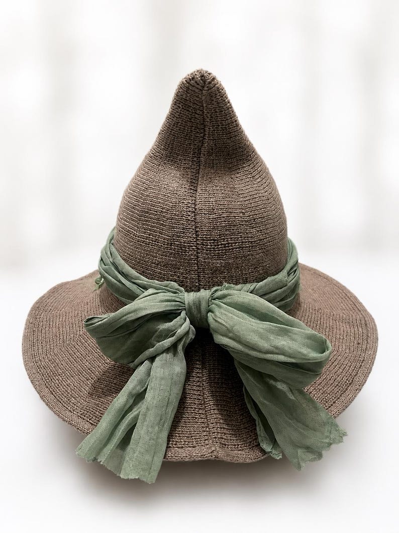 May include: A pointed brown knit hat featuring a wide brim and a sage green fabric ribbon tied into a bow around the base of the crown. The hat is constructed from a textured, solid-colored knit material with a distinct conical shape.