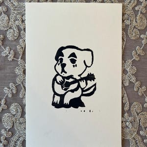 以下が含まれることがあります： ウクレレを演奏する漫画の犬の白黒リノカットプリント。犬は座って楽器を持っています。プリントは白いカードにあり、装飾的なレースの背景があります。