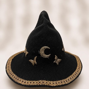 May include: A black pointed hat featuring a wide brim with a metallic gold braided trim. The crown is decorated with small, antique gold-toned metal embellishments, including a crescent moon and several butterflies arranged around the base.