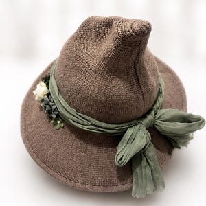May include: A brown knit hat with a pointed crown and a wide brim. The hat is adorned with a sage green fabric ribbon tied in a bow at the front, featuring a cluster of small white and dark green artificial flowers tucked into the side of the band.