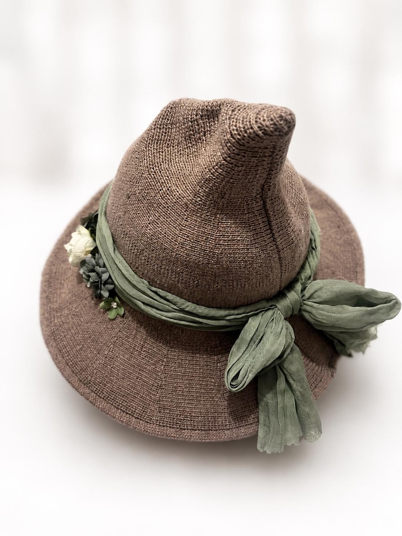 May include: A brown knit hat with a pointed crown and a wide brim. The hat is adorned with a sage green fabric ribbon tied in a bow at the front, featuring a cluster of small white and dark green artificial flowers tucked into the side of the band.