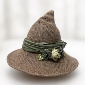 May include: A brown knitted pointed hat with a wide brim. The hat is adorned with a sage green fabric band wrapped around the base, featuring a cluster of small artificial flowers in shades of cream, sage, and dark green on the front side.