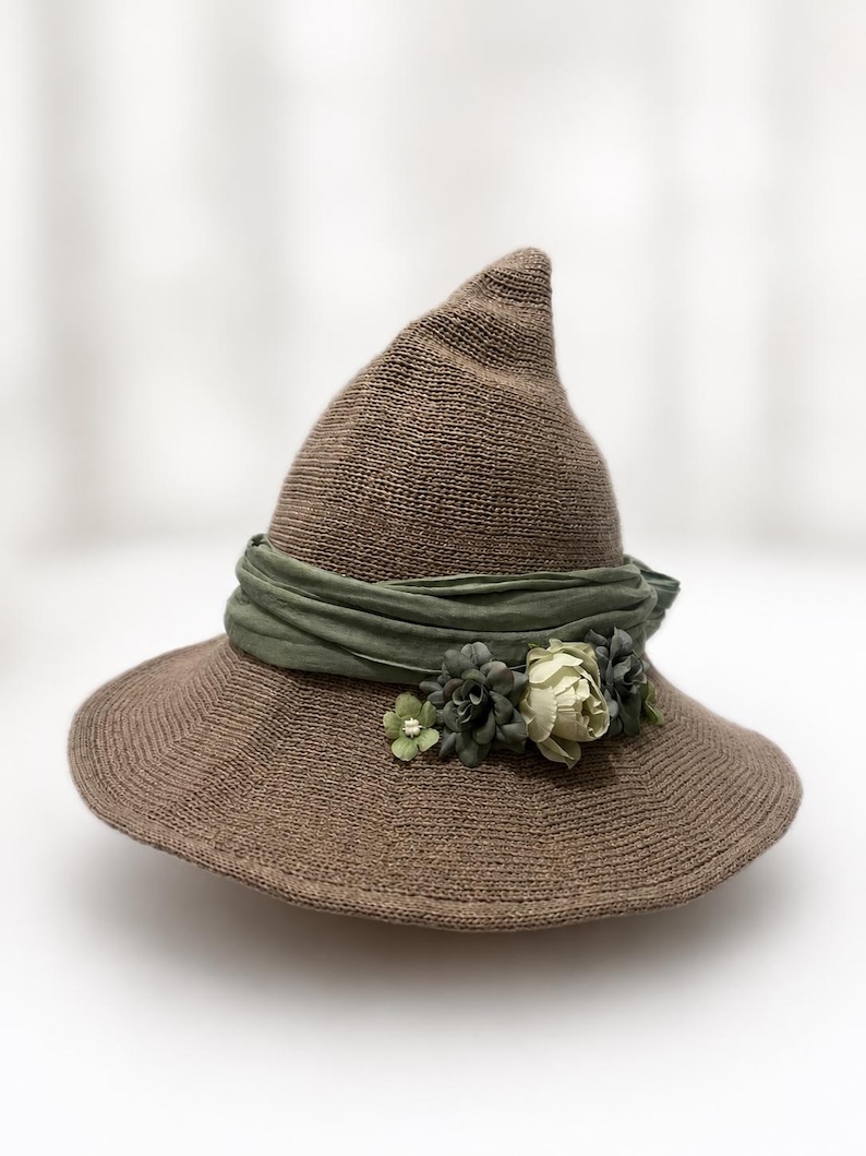 May include: A brown knitted pointed hat with a wide brim. The hat is adorned with a sage green fabric band wrapped around the base, featuring a cluster of small artificial flowers in shades of cream, sage, and dark green on the front side.