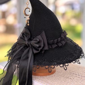 May include: A black pointed hat with a wide brim, featuring a large satin bow, a ruffled band, and a decorative gold-toned charm with a crescent moon and stars hanging from the tip. The brim is finished with a delicate black lace trim.