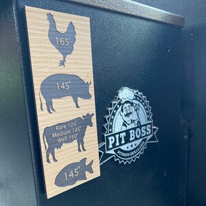 May include: A wooden sign with a black silhouette of a chicken, pig, cow, and fish. The sign has the text "165°", "145°", "Rare 125° Medium 145° Well 160°", and "145°". A silver sticker with the text "PIT BOSS PITBOSS-GRILLS.COM" is on a black surface.