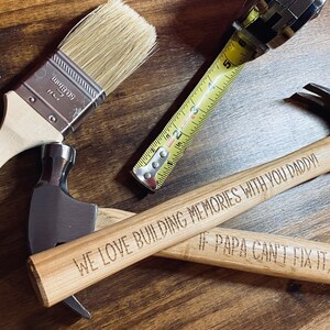 Pode incluir: Dois martelos com cabo de madeira com mensagens gravadas. O primeiro martelo diz "We love building memories with you Daddy!" e o segundo martelo diz "If Papa can't fix it, no one can". Tamb&eacute;m se pode ver um pincel e uma fita m&eacute;trica na imagem.