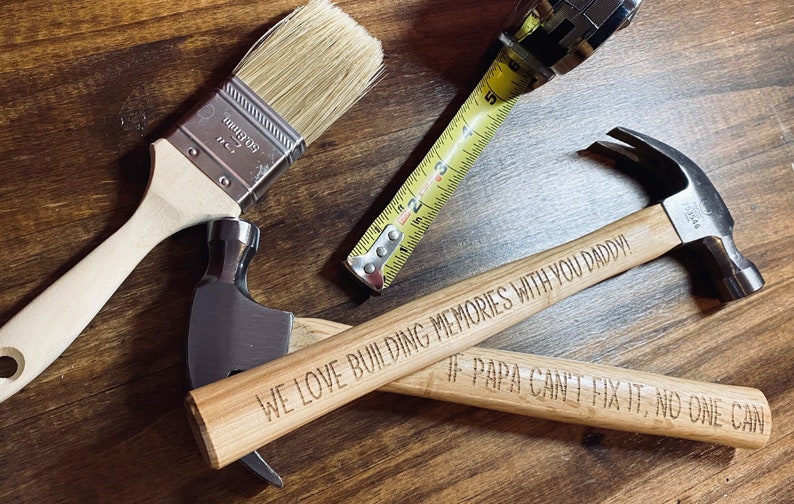 Pode incluir: Dois martelos com cabo de madeira com mensagens gravadas. O primeiro martelo diz "We love building memories with you Daddy!" e o segundo martelo diz "If Papa can't fix it, no one can". Tamb&eacute;m se pode ver um pincel e uma fita m&eacute;trica na imagem.