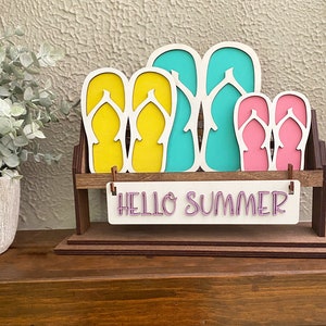 May include: Wooden cutout of flip flops in yellow, blue, and pink colors, arranged on a wooden stand with the text "Hello Summer" on a white sign.