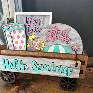 May include: A wooden wagon with wheels, painted in a light gray wash, with the words "Hello Spring" in teal lettering. The wagon is filled with spring-themed decorations, including a sign that says "May Flowers", a sign that says "April Showers", a teal and white striped umbrella, and pink and white polka dot rain boots.