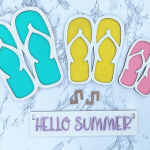 May include: Wooden cutout of flip flops in turquoise, yellow, and pink colors. The cutout is on a white marble background with the text "HELLO SUMMER" on a white sign.