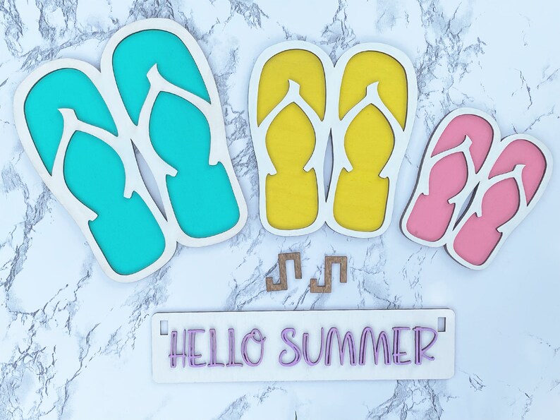 May include: Wooden cutout of flip flops in turquoise, yellow, and pink colors. The cutout is on a white marble background with the text "HELLO SUMMER" on a white sign.