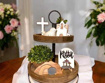 May include: A two-tiered wooden tray with a black metal stand. The top tier features a white church, a white dove, and a wooden cross. The bottom tier has a small potted green plant, a wooden tomb with a rolling stone, and a sign that reads "He is Risen".