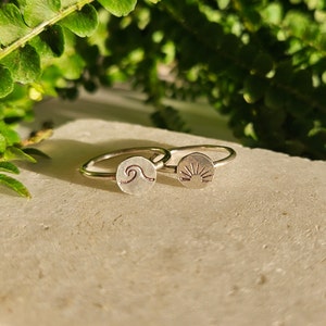 Puede incluir: Dos anillos de plata con un diseño de ola y sol. Los anillos están sobre una superficie de color marrón claro.