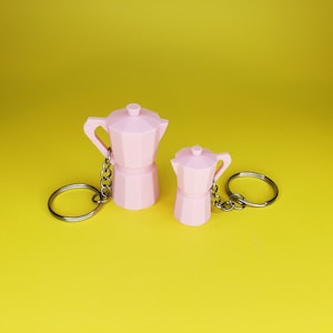 May include: Two pink miniature coffee pot keyrings with silver chains. The larger pot is on the left and the smaller pot is on the right.