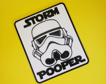 May include: A rectangular white plaque with a black border featuring a raised black graphic of a helmeted character in the center. The word STORM is printed in black above the helmet, and the word POOPER is printed in black below the helmet.