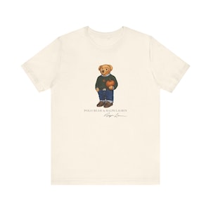 Könnte beinhalten: Ein cremefarbenes Kurzarm-T-Shirt mit einer Grafik eines Teddybären, der einen dunkelgrünen Pullover und eine blaue Jeans trägt und einen Kürbis hält. Der Text POLO BEAR by RALPH LAUREN und eine Unterschrift sind unter der Bärengrafik gedruckt.