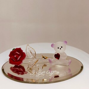 Puede incluir: Figurita decorativa de cristal con una rosa roja, una hoja transparente con detalles dorados y un pequeño oso blanco con un corazón rojo. La figura está sobre una base ovalada espejada con la frase "I Love You".