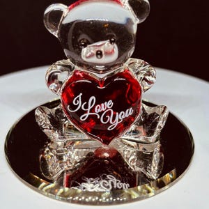Puede incluir: Una figura de oso de peluche de cristal transparente con un corazón rojo. El corazón tiene las palabras "I Love You" en escritura blanca. El oso está sobre una base redonda y espejada. La cabeza del oso es negra y el corazón es rojo intenso.