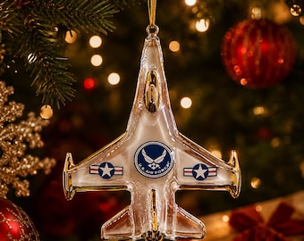Puede incluir: Adorno navideño de avión a reacción de cristal transparente con detalles dorados, colgado de una cinta dorada. El adorno presenta el logotipo de la Fuerza Aérea de EE. UU. y emblemas de estrellas. Cuelga de un árbol de Navidad con bolas rojas y luces.