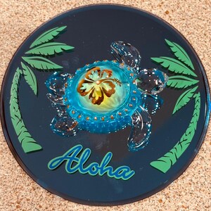 May include: A circular, dark blue decorative item featuring a clear, blue sea turtle design. The turtle has a flower on its shell and is surrounded by green palm trees. The word "Aloha" is written in a teal script font.