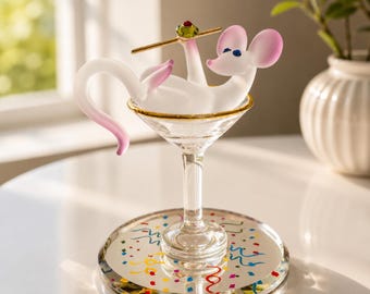 Könnte beinhalten: Eine skurrile Glasfigur einer Maus, die in einem Martini-Glas liegt. Die Maus ist weiß mit rosa Ohren und Schwanz und hält eine goldfarbene Olive. Das Martini-Glas hat einen goldenen Rand und steht auf einem verspiegelten Untersetzer mit bunten Konfetti.