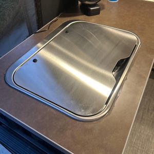 May include: A rectangular stainless steel cover with rounded corners and a recessed handle cutout, integrated into a brown countertop surface. The metal surface features a brushed finish with two small black circular fasteners visible on the top left.