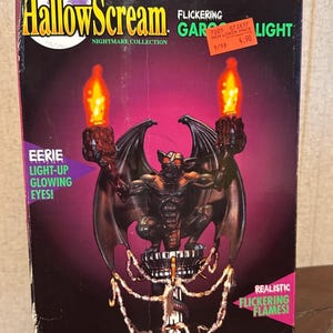 May include: A Halloween-themed box featuring a gargoyle light. The box has the words "HallowScream" and "Nightmare Collection". The gargoyle has light-up glowing eyes and realistic flickering flames. The box is black with purple and red accents.