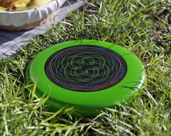 Può includere: Un frisbee verde con un designo astratto colorato al centro. Il frisbee è sdraiato su un letto di erba verde.