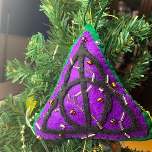 May include: A purple, green, and black felt Christmas tree ornament in the shape of a triangle. The ornament features a black symbol and is decorated with small beads. It hangs from a green ribbon.