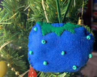 May include: A handmade blue felt Christmas ornament shaped like a blueberry, with green felt leaves and teal bead accents. The ornament has a green velvet loop for hanging and is displayed on a Christmas tree.