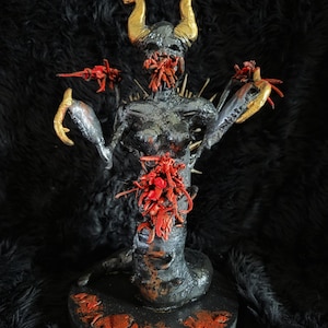 May include: A dark sculpture of a demonic figure with golden horns, metallic gray body, and red accents. The figure has a skeletal face, a spiky torso, and is posed with outstretched arms. It stands on a black, circular base with red paint details.