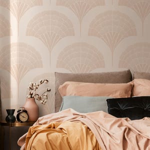May include: A bedroom scene with a bed dressed in peach-toned bedding and pillows. A black velvet cushion and a round, peach-coloured cushion are visible. A small side table holds a vase with cotton stems, a black vase, and a vintage alarm clock. The wallpaper has a fan-like pattern.