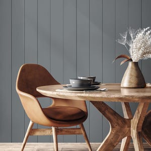 May include: A dining room scene featuring a round wooden table with a natural finish, set with gray plates and bowls. A brown leather chair is positioned at the table. A vase with dried flowers sits on the table. The background is a gray vertical panel wall.