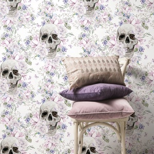 May include: White wallpaper with a floral pattern featuring skulls and butterflies. The wallpaper is behind a white chair with three pillows, one beige, one purple, and one pink.