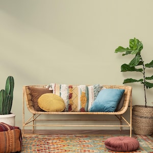 Puede incluir: Un banco de mimbre con varios cojines decorativos en tonos marrones, amarillos y azules. Una alfombra estampada en tonos cálidos se encuentra debajo del banco. Un cactus en maceta y una planta de hojas grandes en una cesta de mimbre a cada lado.
