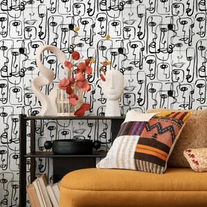 May include: A black and white abstract face pattern wallpaper with a white ceramic sculpture, a glass vase with red flowers, and a black metal shelf with a black bowl and books. The shelf is in front of a yellow couch with a patterned pillow.